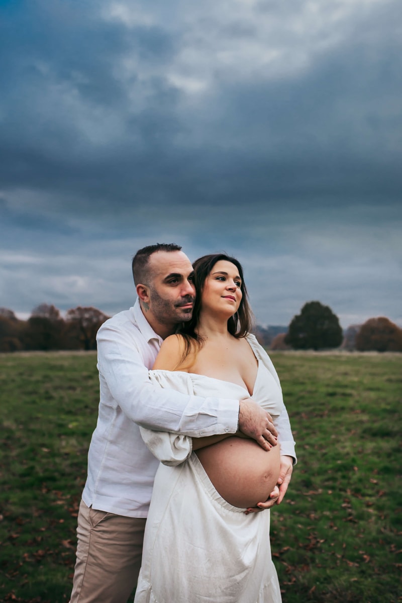 London Family Photographer, man embraces his pregnant partner outdoors, their hands on her belly