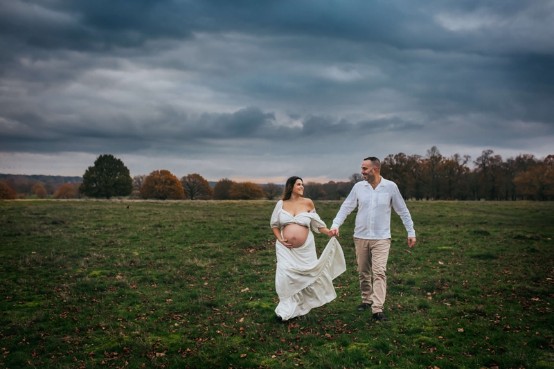 London Family Photographer, a man and expecting woman walk hand in hand through a grassy field